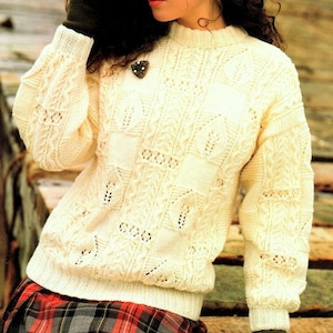 May include: A white knitted jumper with a cable knit pattern and a heart-shaped brooch. The jumper has a round neckline and long sleeves.