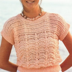 May include: A peach-coloured knitted top with a delicate openwork pattern and short sleeves. The top has a round neckline and a fitted silhouette.