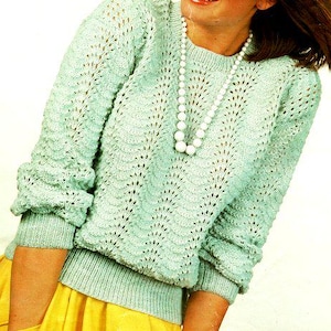 May include: A light blue knitted sweater with a delicate openwork pattern. The sweater has a round neckline and long sleeves. The model is wearing a yellow skirt and a white pearl necklace.