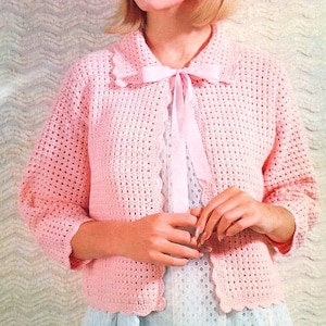 Peut inclure: Un cardigan en crochet rose avec un bord festonné et un nœud papillon. Le cardigan est ouvert à l'avant et a des manches trois quarts.