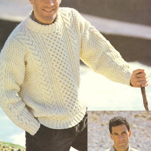 May include: A white cable knit jumper with a crew neck. The jumper has a textured pattern and is worn by a person standing on a rocky beach.