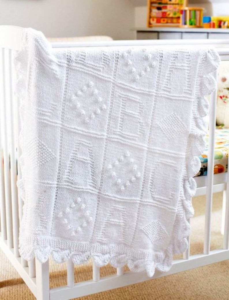 May include: White knitted baby blanket with a scalloped edge and a variety of textured patterns. The blanket is draped over a white crib.
