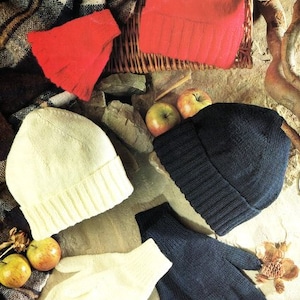 May include: Four knitted hats in different colours: red, white, navy blue, and cream. The hats are arranged on a rustic background with apples and a plaid blanket.