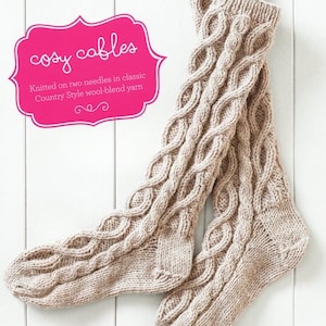 May include: A pair of beige knitted socks with a cable knit pattern. The socks are knitted on two needles in classic Country Style wool-blend yarn. "Cozy Cables" is written on a pink label.