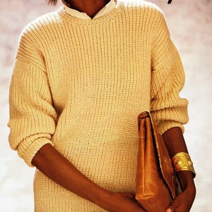 Puede incluir: Un suéter de punto amarillo claro con cuello redondo y mangas largas. El suéter se combina con una camisa blanca con cuello debajo. Se sostiene un clutch de cuero marrón en los brazos. Se usa una pulsera dorada en la muñeca.