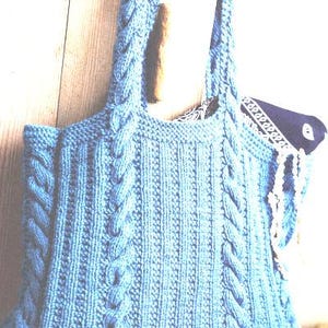 Puede incluir: Un bolso tote tejido de color azul claro con detalles de punto de cable. El bolso tiene dos asas y una forma rectangular. El interior del bolso está forrado con una tela estampada. El bolso está hecho de un hilo suave y texturizado.