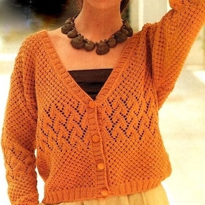 May include: A knitted cardigan with a V-neck and button fastening. The cardigan is orange and has a lacy pattern. The model is wearing a brown top underneath the cardigan.