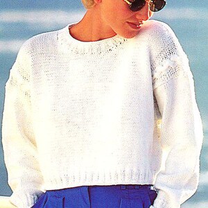May include: A white, crew-neck sweater with textured details on the shoulders and neckline. The sweater is paired with high-waisted, blue pants. The image is set against a blurred background of water and sky.