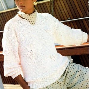 Puede incluir: Un suéter blanco de punto con cuello en V y diseño de punto de cable. El suéter tiene un patrón floral y se usa con una falda de lunares verdes y blancos.