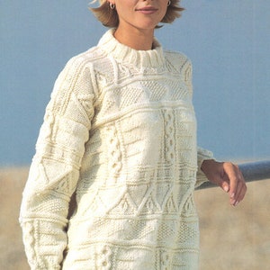 May include: Cream-colored, long-sleeved sweater with intricate cable-knit and textured patterns. The sweater has a high, ribbed collar and a slightly oversized fit. The text "THE SPINNING WHEEL 88 MARKET STREET EARLESTOWN" is visible in the background.