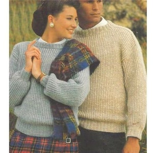 May include: A couple wearing knitted sweaters. The woman is wearing a light blue sweater with a plaid kilt and a scarf. The man is wearing a beige sweater.