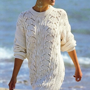 Puede incluir: Un suéter blanco de ganchillo con un delicado patrón de calado. El suéter tiene un cuello redondo y mangas largas. La modelo lleva el suéter en una playa.