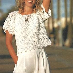 Puede incluir: Un top blanco de crochet con mangas cortas y un dobladillo festoneado. El top tiene un diseño de tejido suelto y abierto. La persona lleva pantalones cortos blancos. El fondo está borroso.