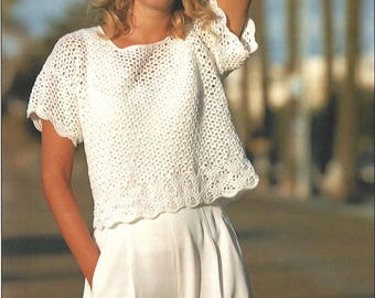 May include: A white crochet top with short sleeves and a scalloped hem. The top has a loose, open weave design. The model is wearing white shorts. The background is blurred.
