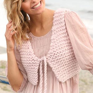 May include: A light pink, chunky knit shrug with a tie closure. The shrug is worn over a light pink blouse with long sleeves. The knitwear has a textured appearance, and the tie is made of a twisted cord.