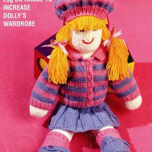Puede incluir: Una muñeca de punto con cabello rubio, que lleva un suéter a rayas rosa y morado, una falda morada y botas moradas. La muñeca sostiene un pequeño gorro rosa con pompón. El texto "CHOOSE OTHER COLOURS IN OUR 25g DK RANGE TO INCREASE DOLLY'S WARDROBE" es visible en el fondo.