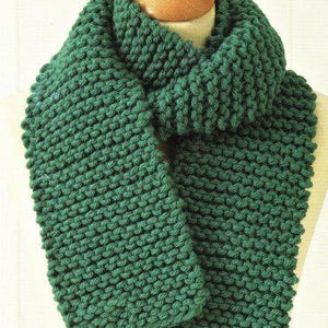 Könnte beinhalten: Ein grüner Strick Schal mit einer grobmaschigen Strickstruktur. Der Schal ist in der Mitte gefaltet und über den Hals einer Schaufensterpuppe gelegt.