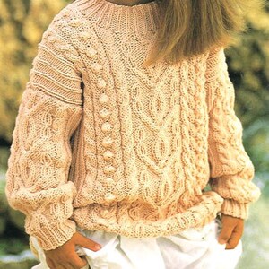 Könnte beinhalten: Ein cremefarbener Strickpullover mit einem Zopfmuster und einem runden Ausschnitt. Der Pullover hat eine strukturierte Oberfläche und ist aus einem weichen, warmen Material gefertigt.