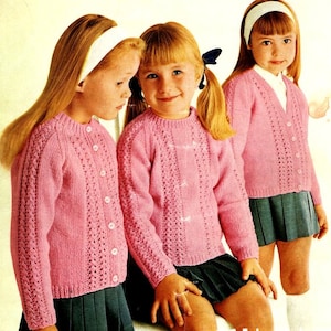 May include: Three girls wearing pink knitted cardigans with button fastenings. The cardigans feature a delicate lace pattern. The girls are wearing matching dark blue skirts.