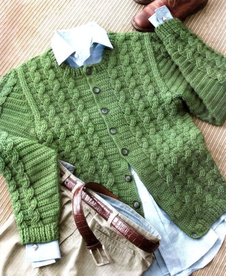 Op de afbeelding: Een groene gehaakte cardigan met een knoopsluiting en lange mouwen. De cardigan is gemaakt met een kabelsteek.
