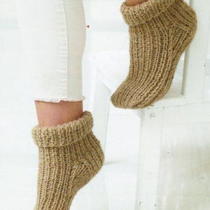 Puede incluir: Primer plano de dos calcetines de punto beige con textura acanalada. Los calcetines tienen un puño doblado y se usan con pantalones blancos. Los calcetines parecen hechos a mano y son de un estilo cálido y acogedor.