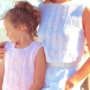 May include: A mother and child wearing matching sleeveless knitted tops. The mother's top is white, and the child's is pink. Both tops have a textured, openwork design. They are standing near the sea.