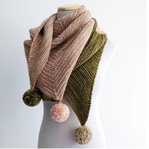 May include: A knitted shawl in shades of pink and olive green, displayed on a mannequin. The shawl features a triangular design with three pom-poms in coordinating colors. The knitted texture is clearly visible, showcasing the craftsmanship of the piece.