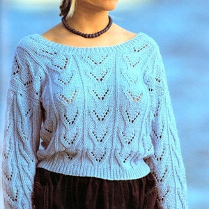 Può includere: Un maglione di lana blu chiaro con un motivo a pizzo. Il maglione ha un collo rotondo e maniche lunghe. La modella indossa una gonna nera.