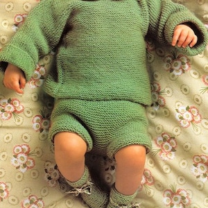 May include: A baby wearing a green knitted sweater and matching shorts with a white crocheted collar. The baby is lying on a floral patterned blanket.