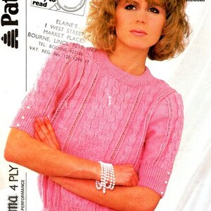 May include: A pink knitted sweater with a cable knit pattern and pearl buttons on the sleeves. The sweater is shown on a model with short, curly blonde hair. The text on the image reads "Diploma 4 Ply High Performance" and "Large Print easy to read".