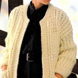 May include: A white crocheted cardigan with a cable knit pattern and pockets. The cardigan is open in the front and has a black shirt underneath.