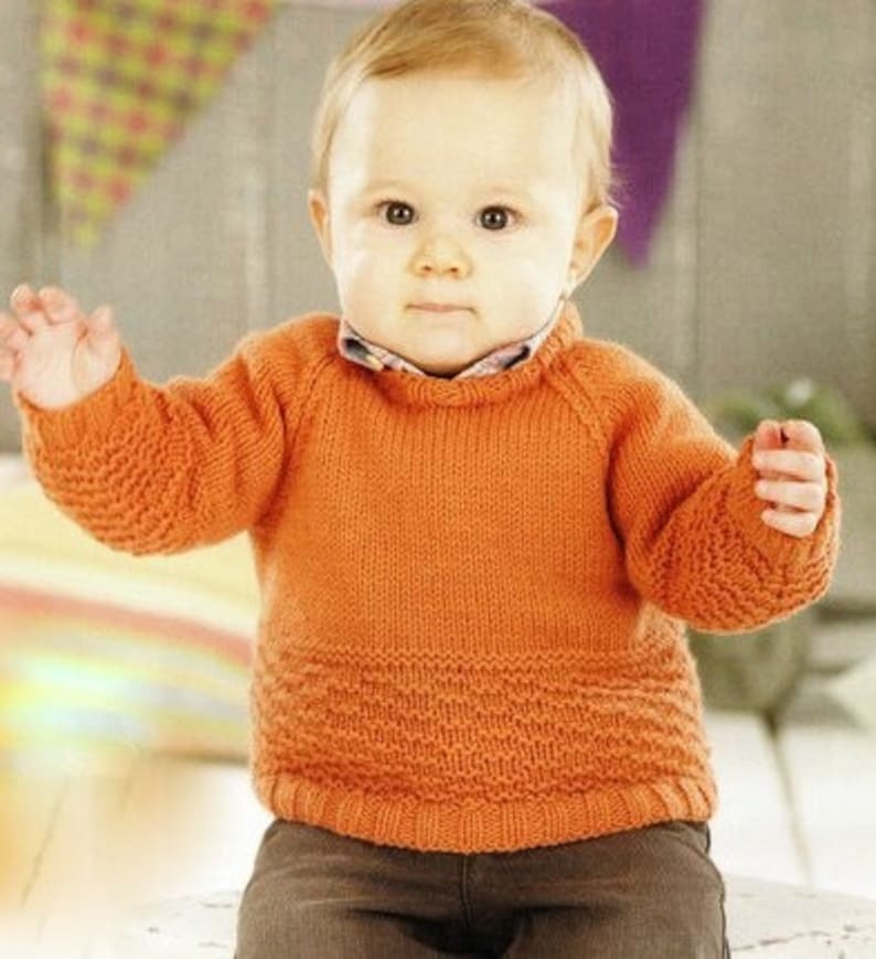 Peut inclure: Un pull orange tricot&eacute; avec une section inf&eacute;rieure textur&eacute;e et des poignets c&ocirc;tel&eacute;s. Le pull a une encolure ronde et des manches longues. Le b&eacute;b&eacute; porte un pantalon marron. L'arri&egrave;re-plan comprend une banni&egrave;re color&eacute;e.