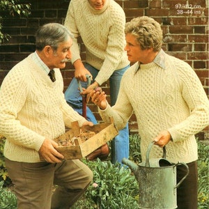 May include: Three men wearing cream-colored Aran knit sweaters. The sweaters have a cable knit pattern. The men are standing in a garden setting. The text "Aran 97-112 cm 38-44 ins" is visible in the upper right corner of the image.