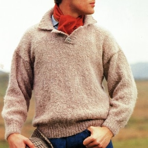 Puede incluir: Un hombre lleva un suéter marrón claro con un cierre de botón único en el cuello. Lleva una bufanda roja alrededor del cuello y pantalones vaqueros azules. Sostiene un sombrero de cuadros marrones y negros en su mano izquierda.