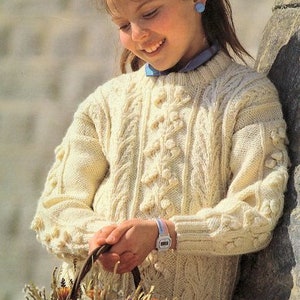 May include: A young girl wearing a cream-colored cable knit sweater with a blue bow in her hair holds a basket of dried flowers.