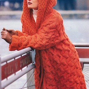 May include: A long, orange knitted cardigan with a hood. The cardigan has a cable knit pattern and is worn by a person with long brown hair.