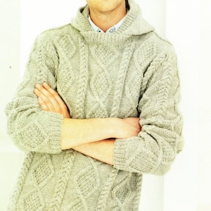 Puede incluir: Un suéter de punto gris claro con un patrón de punto de cable y una capucha. El suéter tiene un ajuste holgado y un aspecto ligeramente de gran tamaño.