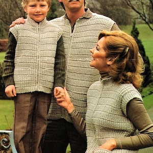 May include: A family of three wearing knitted vests. The vests are beige and have a zipper closure. The vests are sleeveless and have a ribbed texture. The family is standing in front of a green grassy area.