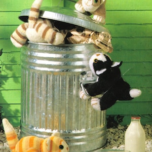 May include: Four knitted cat toys in a silver metal trash can. One cat is hanging from the edge of the can, one is inside the can, and two are outside the can. There is a glass milk bottle and a glass milk jug on the ground.