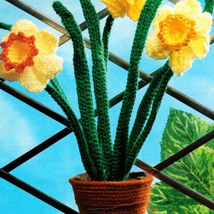 May include: A crocheted daffodil plant with three yellow flowers in a brown crocheted pot. The plant is against a blue and white background.