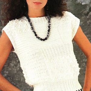 May include: A white knitted sleeveless top with a round neckline and a beaded necklace. The top has a textured pattern and a ribbed hem.