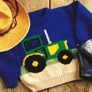 Puede incluir: Un suéter de punto azul con un diseño de tractor verde y amarillo. El suéter tiene un ribete blanco alrededor del cuello y un fondo de color crema.