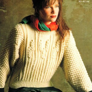 May include: A close-up of a woman wearing a cream-colored Aran knit sweater with a green and red scarf. The sweater has a cable knit pattern and is 30-40 inches in length.
