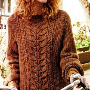 May include: A woman wearing a brown cable knit sweater with a leaf pattern. She is holding a bicycle handlebar.
