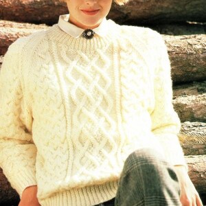 May include: A white cable knit sweater with a round neckline. The sweater has a detailed cable knit pattern and is worn by a person wearing a brown and white patterned hat.