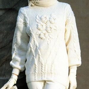 Op de afbeelding: Witte gebreide trui met een kabelbreipatroon en een bloemenmotief op de voorkant. De trui heeft een rolkraag en lange mouwen.