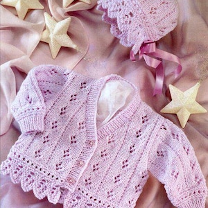 May include: A light pink, knitted baby cardigan and bonnet set. The cardigan has a scalloped edge, button closure, and decorative openwork. The bonnet features a matching scalloped brim and ribbon ties. The set is displayed on a pink fabric with star-shaped decorations.