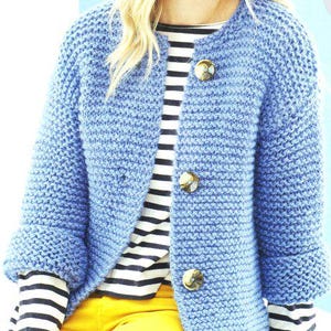 以下が含まれることがあります： 大きな丸いボタンが付いた、水色のニットカーディガン。カーディガンは、白と黒のストライプのシャツと黄色のパンツの上に着用されています。袖口は手首で折り返されています。