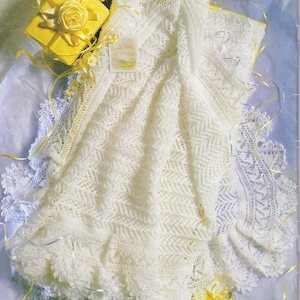 May include: A white crocheted baby blanket with a delicate lace border. The blanket is laid out on a light blue surface with yellow gift boxes.