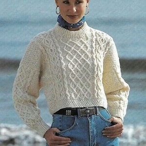 Könnte beinhalten: Ein weißer Strickpullover mit Zopfmuster, Rundhalsausschnitt und langen Ärmeln. Der Pullover ist kurz geschnitten und wird mit einem blauen Jeansrock getragen.